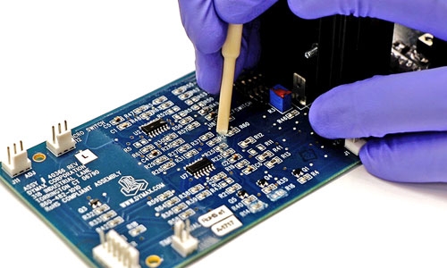 A lab tech uses a plastic tool to remove a cured conformal coating on a printed circuit board.