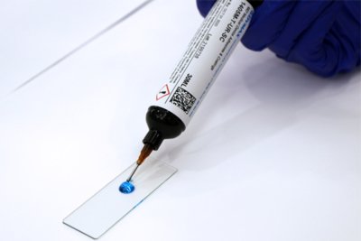 El adhesivo para dispositivos médicos 1405M-T-UR-SC MD® de Dymax dispensándose en azul sobre portaobjetos de plástico